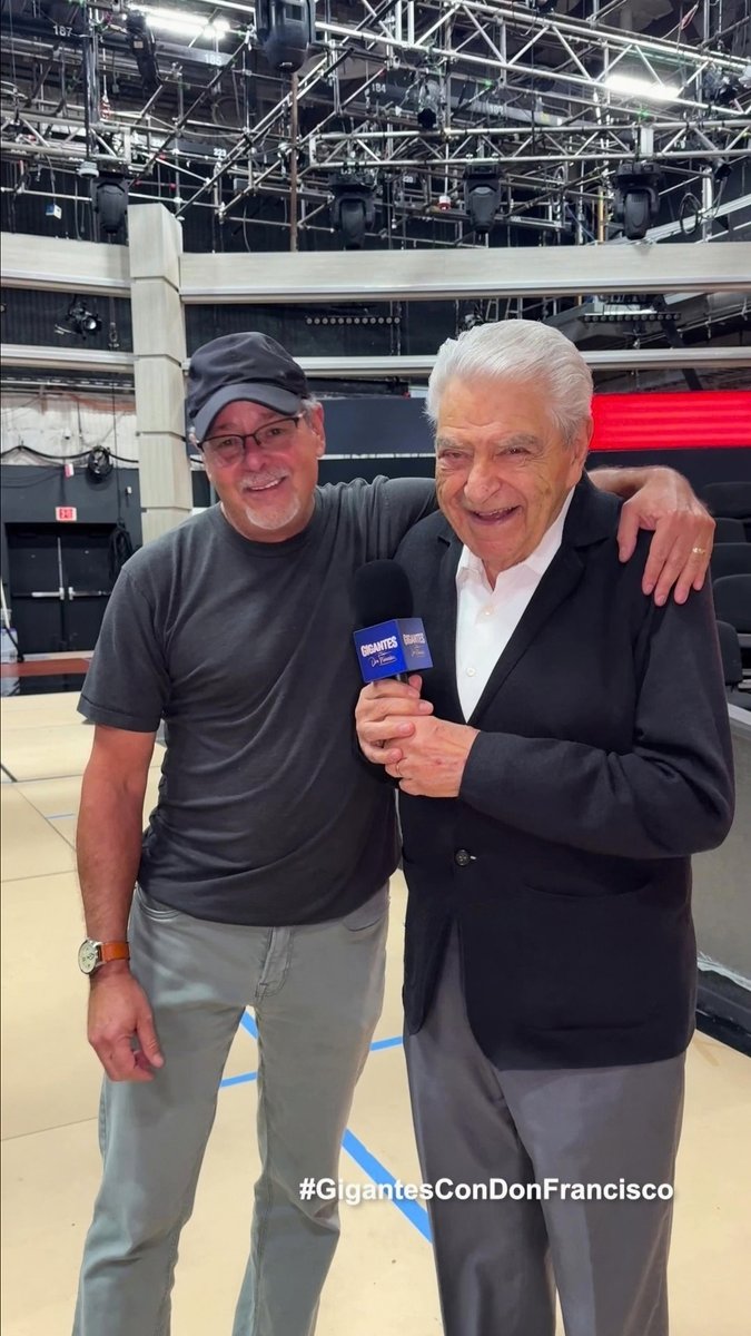 Two men pose together in what appears to be a TV studio; one wears a cap and gray shirt, while the older man in a suit holds a microphone labeled 'GIGANTES.' The caption says '#GigantesConDonFrancisco'.