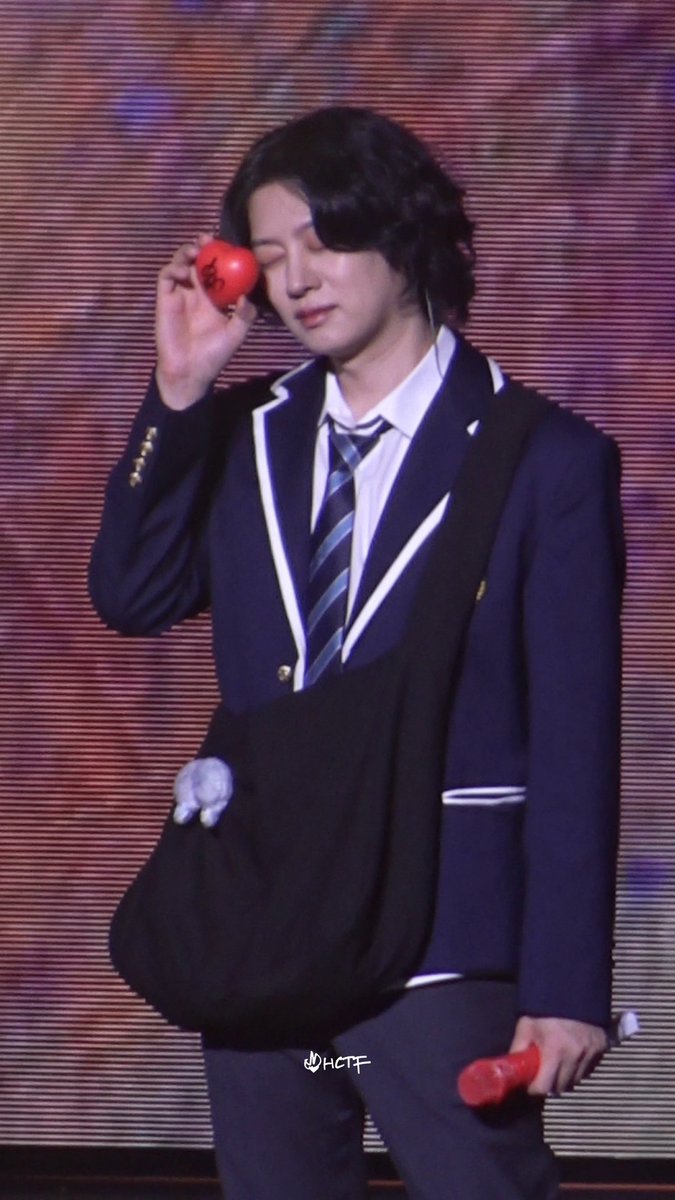 A performer on stage in a navy school uniform-style suit and a crossbody bag, holding a red object near his face and a red microphone in his other hand.
