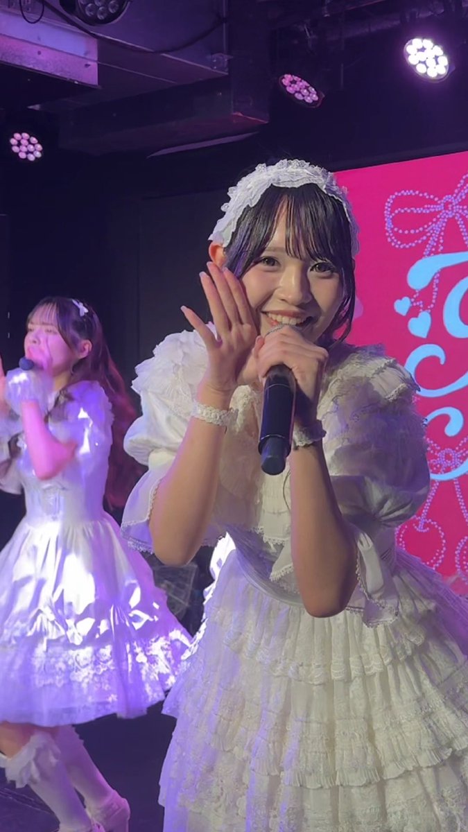 A smiling performer in a frilly white dress and headband holds a microphone on stage, with another performer in a similar white outfit visible in the background.