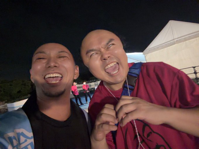 Two young men take a cheerful nighttime selfie, both laughing with their eyes scrunched shut, one wearing a black shirt and the other a red garment, with white tents visible in the background.