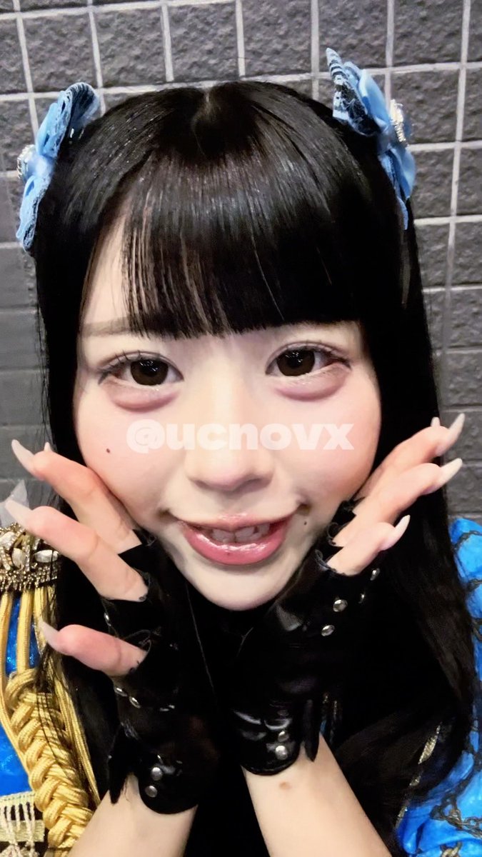 A close-up selfie of a young woman with dark hair, blue hair accessories, and long fingernails posing in front of a gray brick wall, with a watermark reading "@ucnovx".