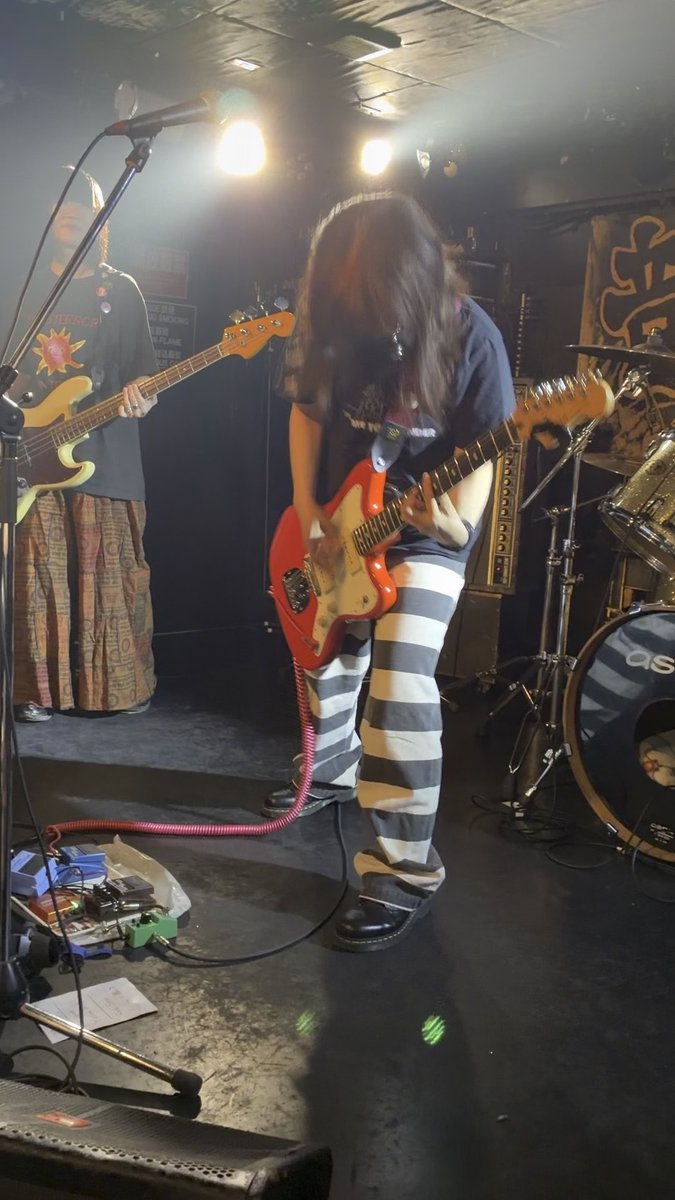 A guitarist performing on stage with long hair covering their face, playing a red Fender Jaguar while wearing a dark t-shirt and striped pants.