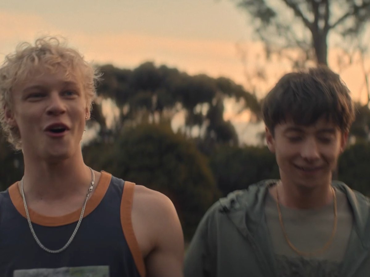 Two young men stand outdoors at sunset, one with curly blonde hair in a dark tank top with orange trim and a silver chain, the other with brown hair in an olive hoodie and gold chain, both smiling in a warm, golden-lit scene that fits the dreamy "Self Control" soundtrack pairing with the horror film *Leviticus (2026)*.