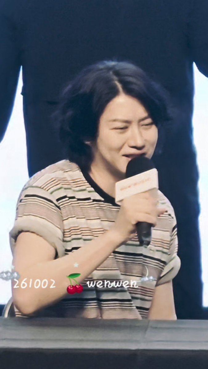 A man with shoulder-length black hair in a striped shirt holds a microphone and smiles, with text overlays '261002', a cherry emoji, and 'wenwen'.