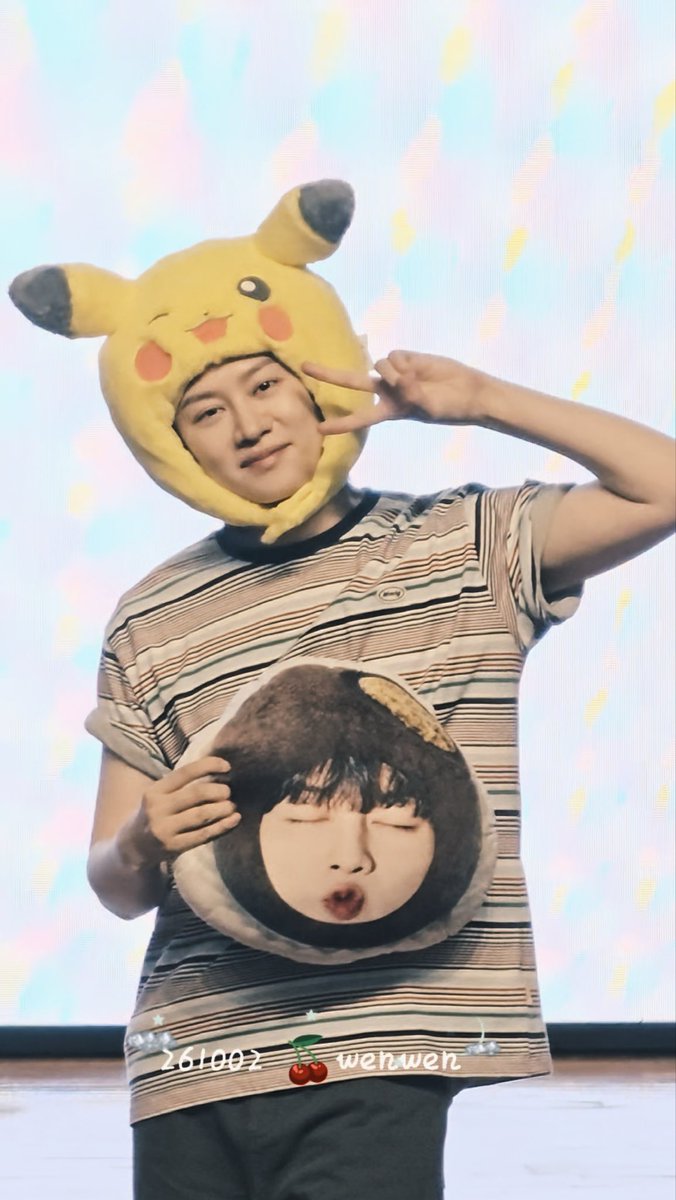 A young man wearing a striped shirt and a Pikachu hat, holding a circular pillow with a person's face on it and making a peace sign.