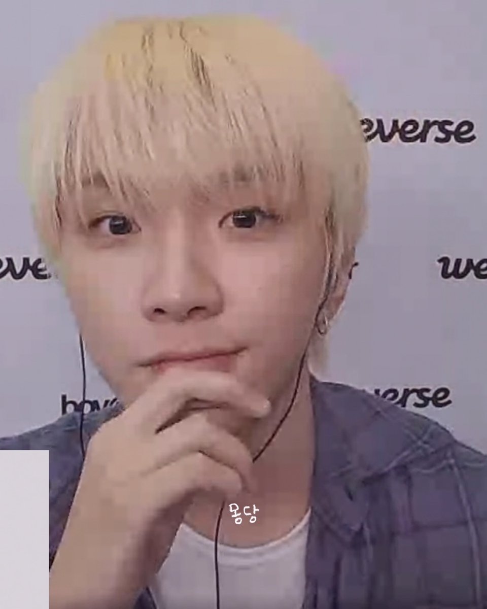A close-up selfie of a young man with blonde hair, wearing a denim jacket and earphones, posing thoughtfully with his hand near his mouth against a 'weverse' backdrop.