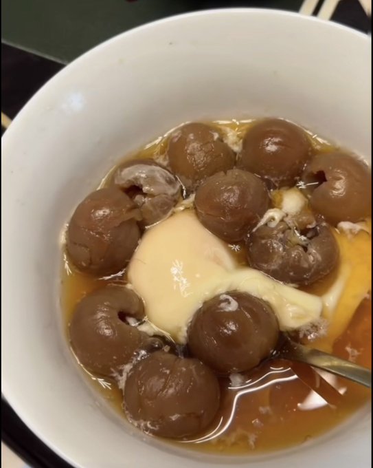 A white bowl containing dark brown balls (possibly meatballs or dumplings) in a brown broth, topped with a melted yellowish substance that resembles melted cheese or butter.