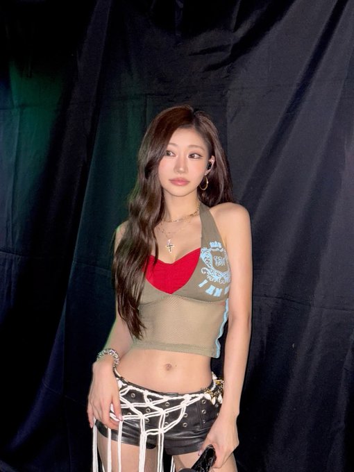 A woman wearing a red bra under a sheer olive crop top and a distressed black skirt with chain details poses against a black backdrop.
