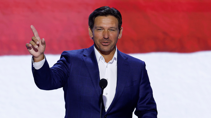 Florida Governor Ron DeSantis in a blue suit pointing upward while speaking at a podium.