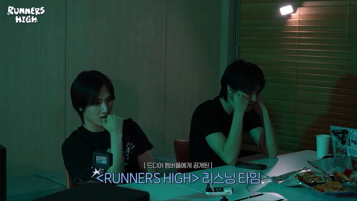 Two men sit at a table in a dimly lit room with a green hue during what appears to be a listening session for the group RUNNERS HIGH, with Korean subtitles on screen.