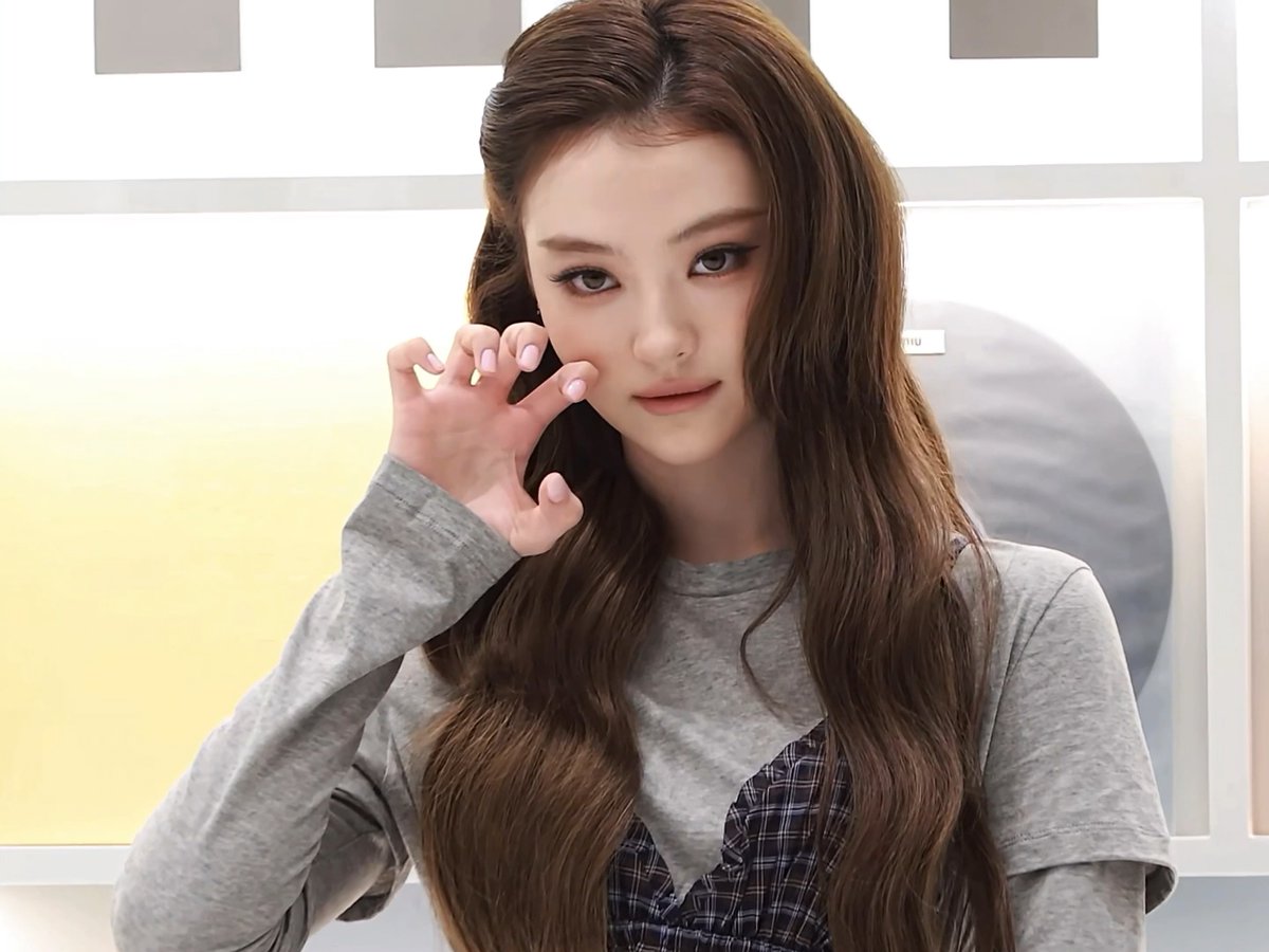 A woman with long wavy brown hair wearing a gray long-sleeve top over a plaid garment poses with her hand near her face in front of a white background.