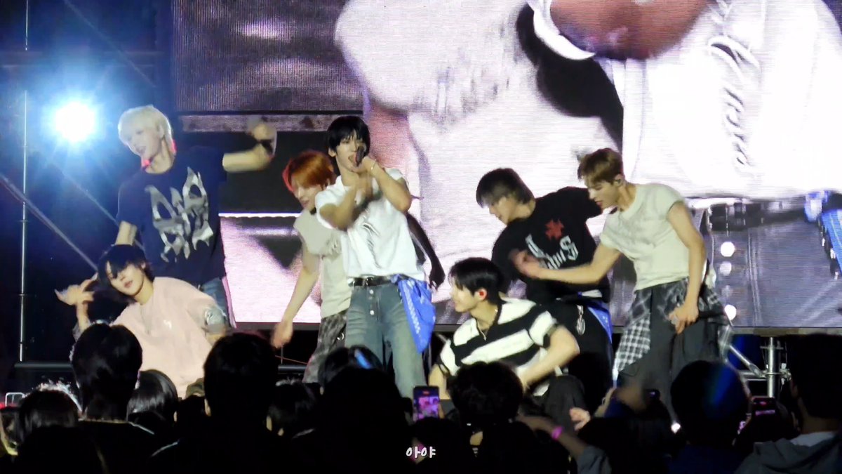 A K-pop group performs energetically on stage in front of a large screen and audience members, with the watermark '아야' visible.
