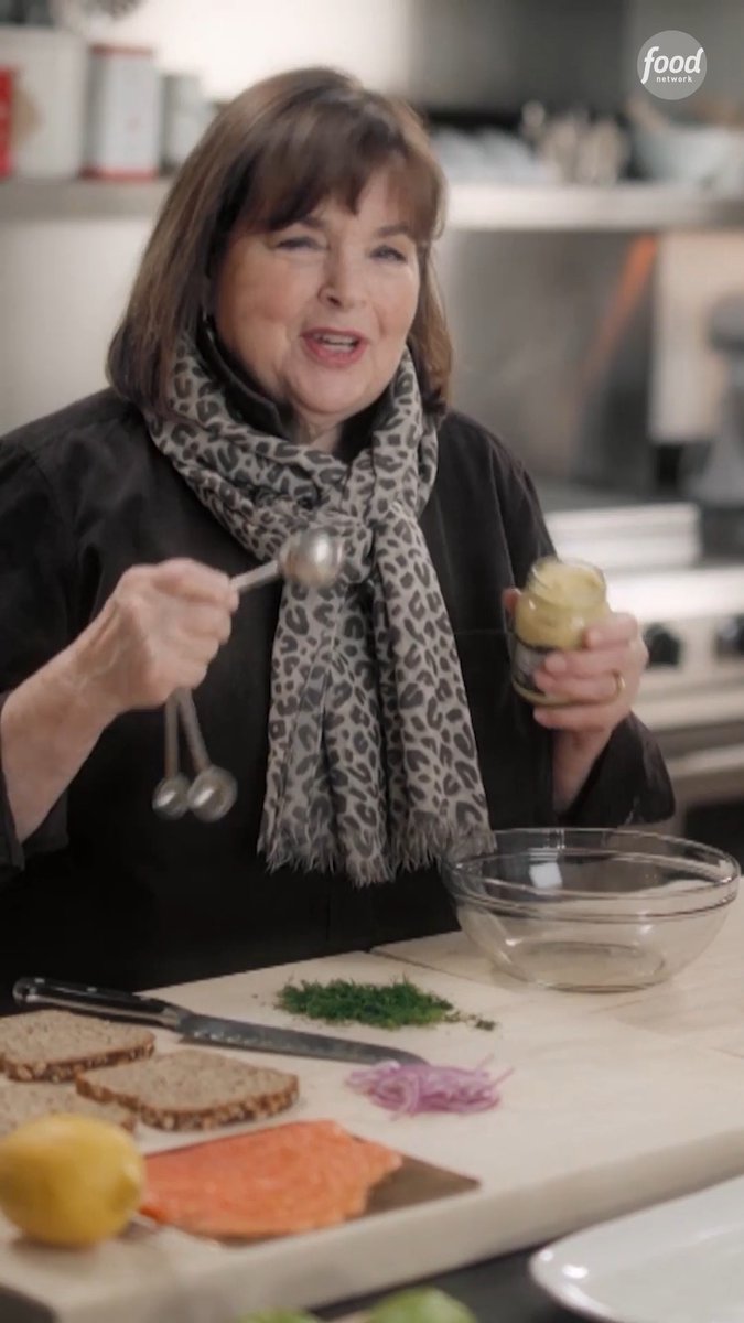 Ina Garten stands in a kitchen holding a jar of Dijon mustard and measuring spoons, with prepared ingredients including salmon, red onion, dill, bread, and lemon on the cutting board, branded with the 'food network' logo.