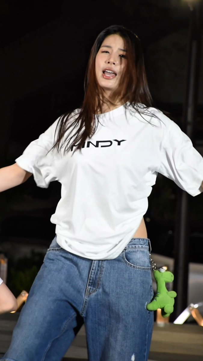 A woman performs on stage at night, wearing a white t-shirt with 'WINDY' printed on it and baggy jeans, with a green plush keychain hanging from her pocket.