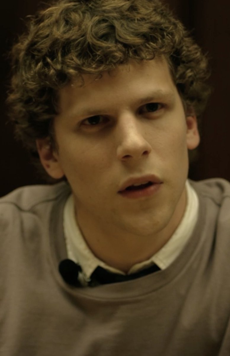 A young man with curly blonde hair and a contemplative expression sits in a dimly lit setting, wearing a gray sweatshirt over a collared shirt with a small lapel microphone clipped to his neckline—visually reinforcing the caption "He's wired in" by showing he is literally fitted with audio equipment for a recording or interview.