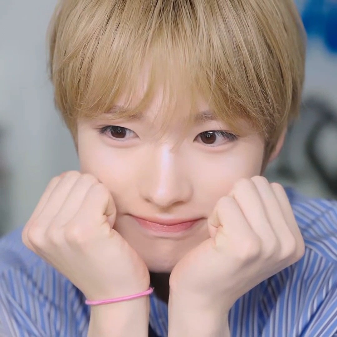 A close-up portrait of a young man with blonde hair resting his face on his hands, wearing a blue striped shirt.