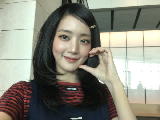 A selfie of a smiling woman with long dark hair wearing a red striped shirt under a dark apron, posing indoors.