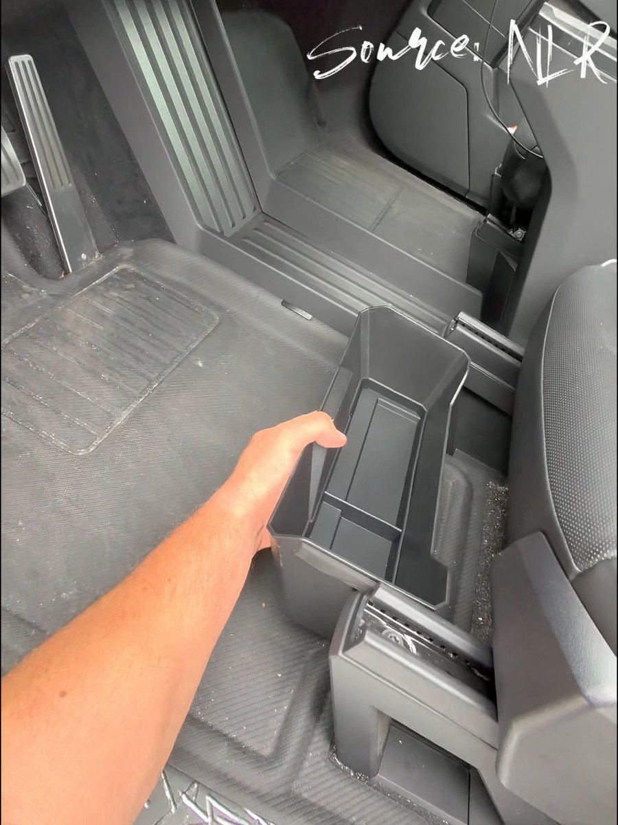 A first-person perspective shows a hand holding a black plastic storage bin above the floor of a vehicle, near the center console, with 'Source: NIK' written in white script in the upper right corner.