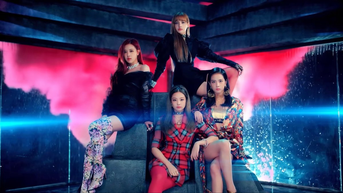 Four women pose together in a stylized promotional photo with pink and blue neon lighting, wearing black outfits and statement jewelry.
