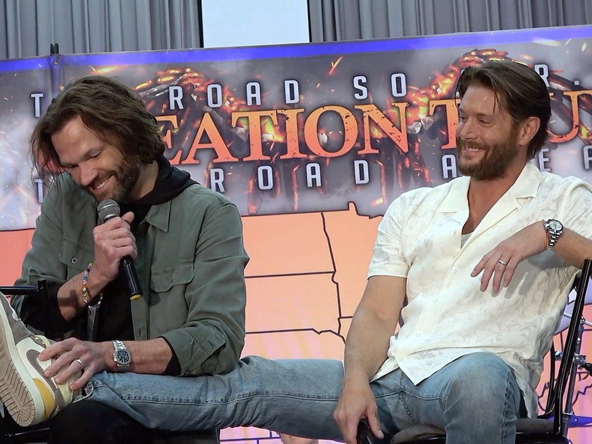 Two bearded men sit at a panel event with a 'STATION 19' backdrop featuring fire imagery, one holding a microphone and smiling, while the other in a white shirt laughs.