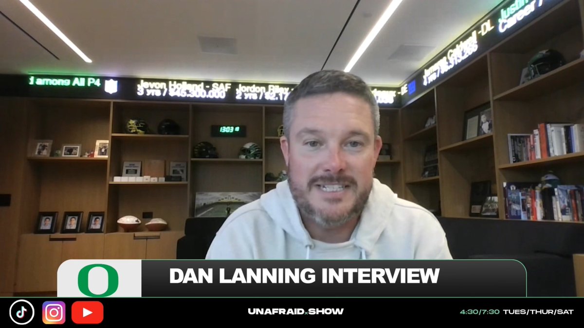 Dan Lanning appears on a video interview set for the "Unafraid Show," as indicated by the Oregon "O" logo and "DAN LANNING INTERVIEW" lower-third banner, while discussing Oregon's cinematic game recaps in the office featuring football memorabilia and scrolling player stat tickers overhead.