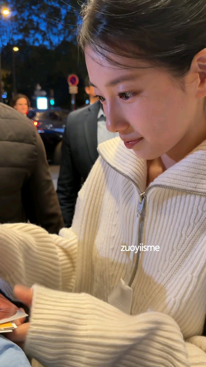 A close-up night shot of a young woman in a cream-colored ribbed zip-up sweater looking down, with a watermark reading 'zuoyiisme' across the image.