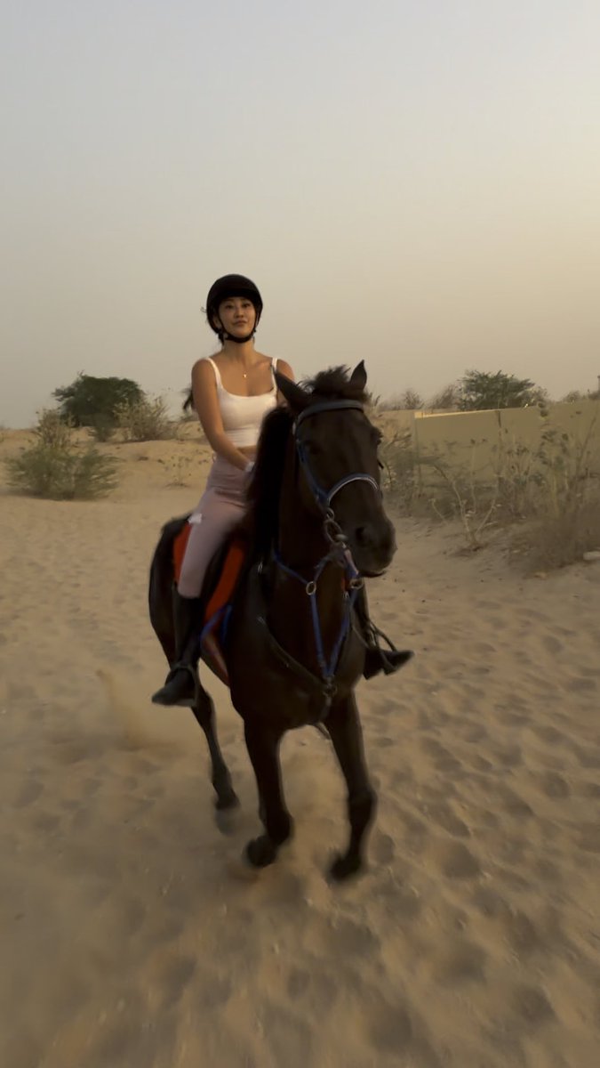 A young woman wearing a black helmet and white tank top rides a dark horse through a sandy desert landscape at sunset.
