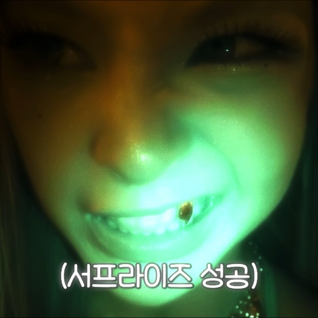 A close-up selfie shows a person's lower face dramatically lit by green light, smiling to reveal a small fang between their teeth. Korean text at the bottom reads "사프라이즈 성공" (Surprise success), playfully captioning the vampire Halloween look the poster requested.