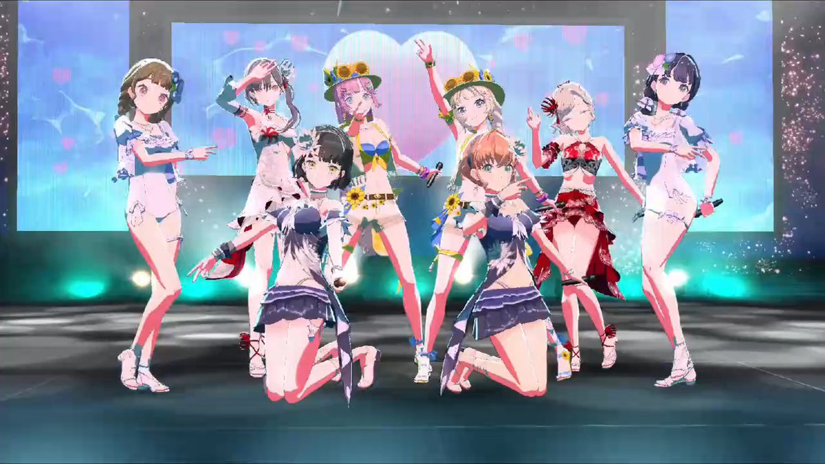 A group of anime-style female characters in colorful, decorative outfits pose and dance on a brightly lit stage with sparkles and a heart design on the screen behind them.