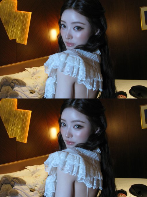 A young woman with long dark hair wearing a white ruffled top poses for a mirror photo, looking over her shoulder with a teddy bear visible on the bed behind her.