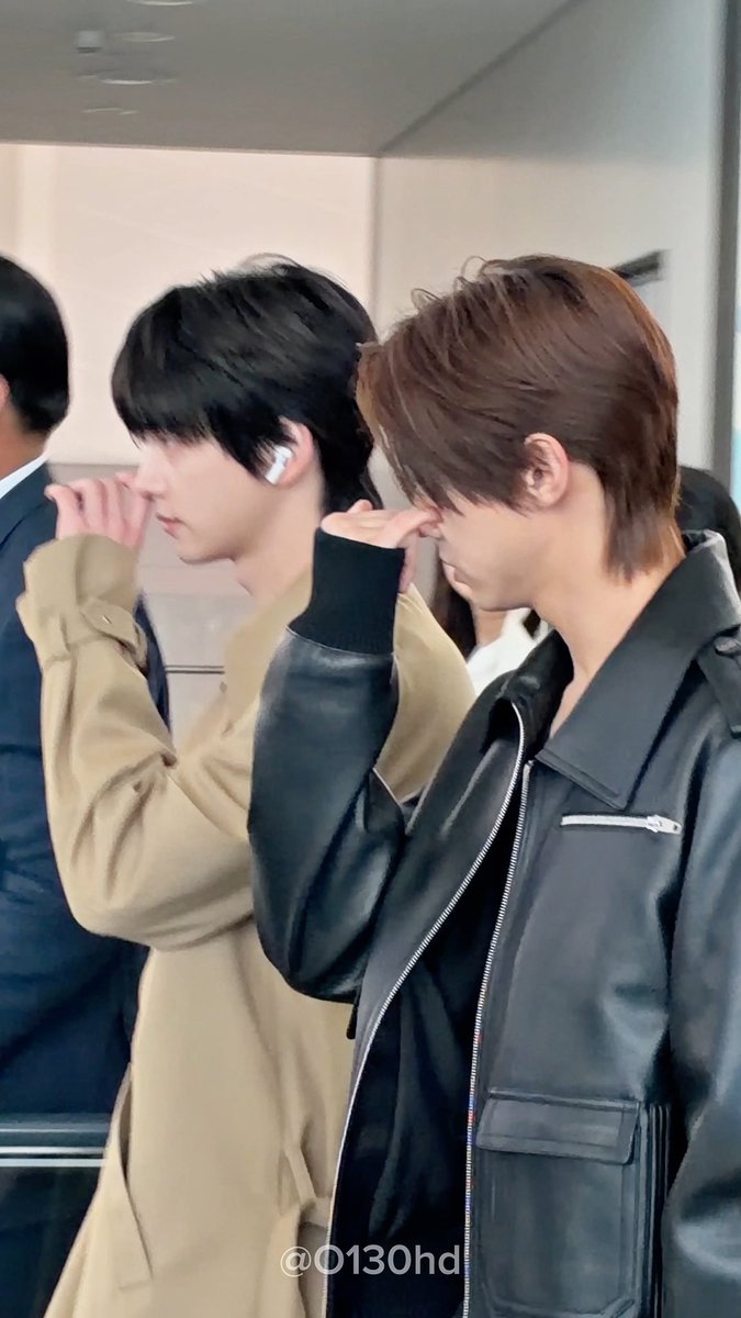 Two young men walk side by side indoors; one wears a tan jacket and a wireless earbud, while the other wears a black leather jacket, both touching their faces, with a watermark '@0130hd'.