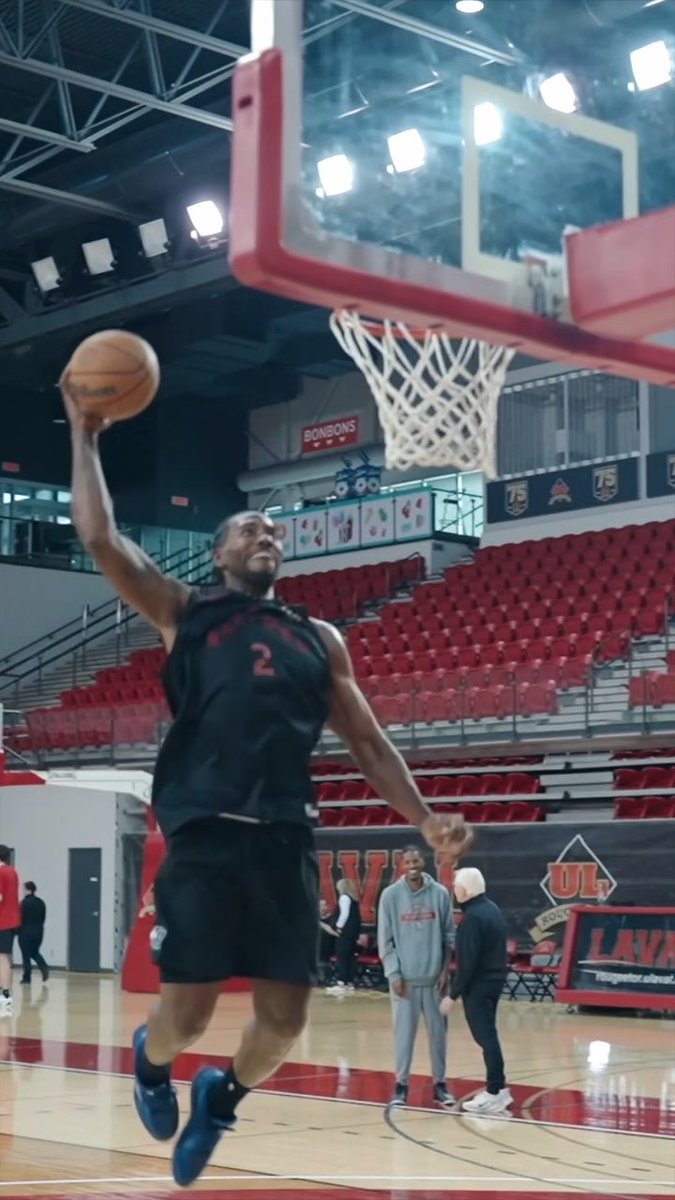 Basketball player #2 leaps toward the hoop for a dunk during a practice session at a UL (University of Louisiana at Lafayette) arena, with "BONJOURS" visible on signage and "75" on banners in the background, illustrating a lighthearted Kawhi vs. Darko post-practice moment.