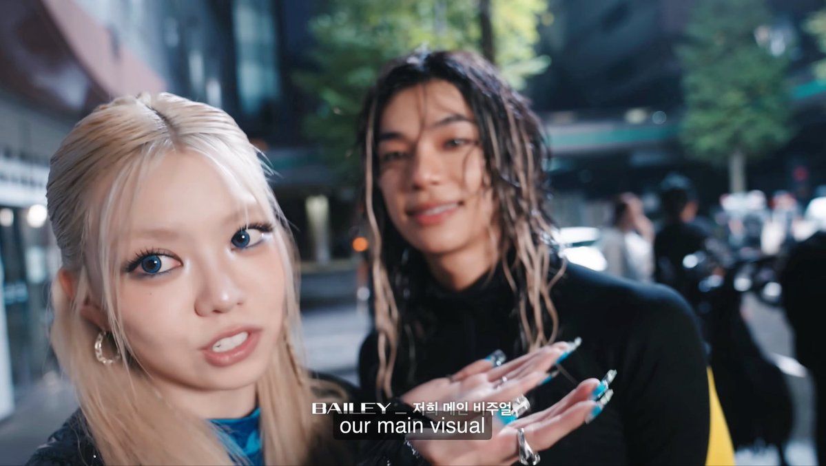 Two women pose together in a nighttime outdoor setting, likely in South Korea; the woman on the left has blonde hair and wears a black jacket, while the woman on the right has long wavy brown hair and a black top. On-screen text in Korean and English reads 'BAILEY _ 저희 메인 비주얼' and 'our main visual'.