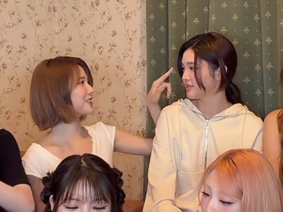 Two young women interact playfully in front of a floral patterned wall, with one in a white top and the other in a cream hoodie pointing near her face.