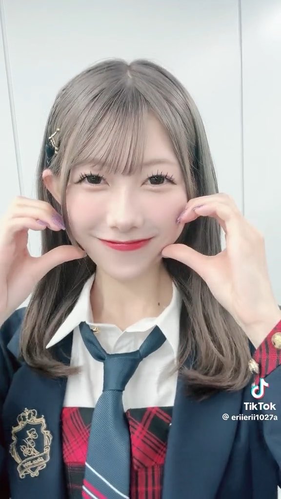 A smiling young woman in a school uniform with a plaid skirt and tie, forming a heart shape with her hands near her face, from a TikTok by erierii1027a.