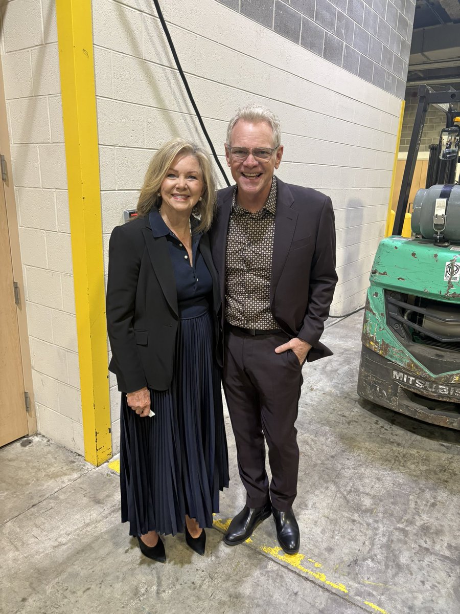 A smiling woman and man pose together in an industrial indoor space. A forklift is parked beside them, and there is text on the machinery.