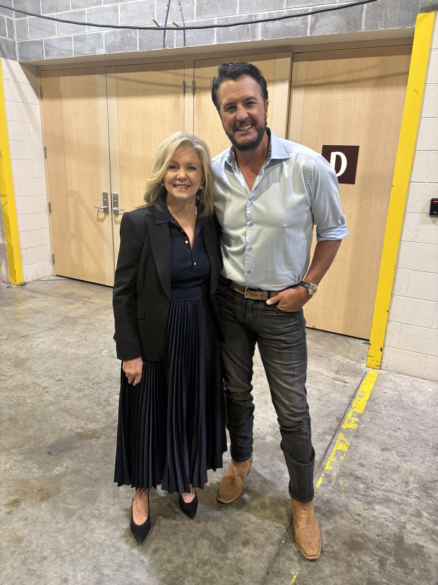 A smiling woman and man pose together in an indoor hallway in front of wooden double doors. A letter sign is visible on one of the doors.