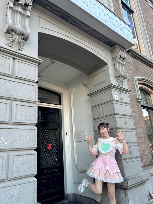 A young woman in a pink and white frilly dress posing with peace signs in front of a classical-style building entrance with ornate stone carvings and a dark wooden door.