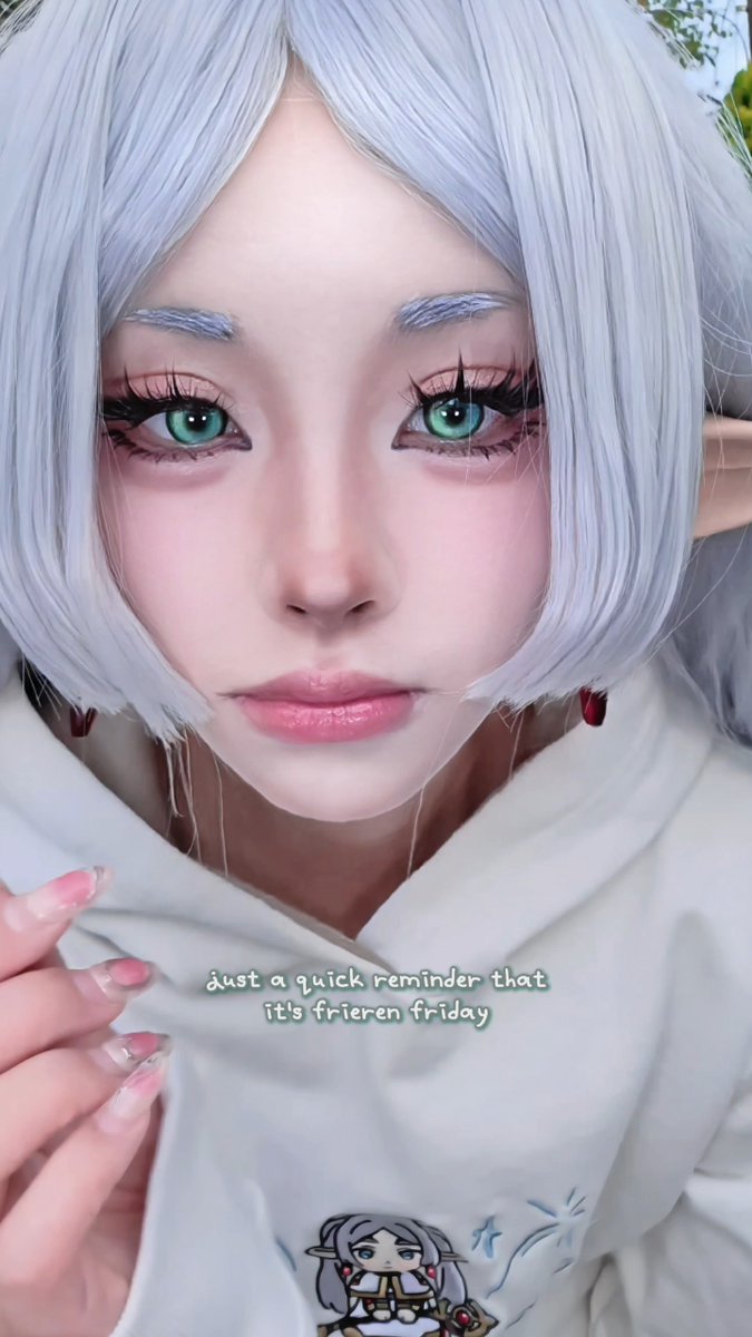 A cosplayer with silver hair, green eyes, and pointed ears dressed as the character Frieren, wearing a white hoodie with a small Frieren graphic, with text reading 'just a quick reminder that it's frieren friday'.