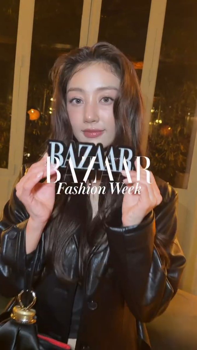 A young woman in a black leather jacket holding up a phone or card, overlaid with 'BAZAAR Fashion Week' text, photographed at night near glass doors.