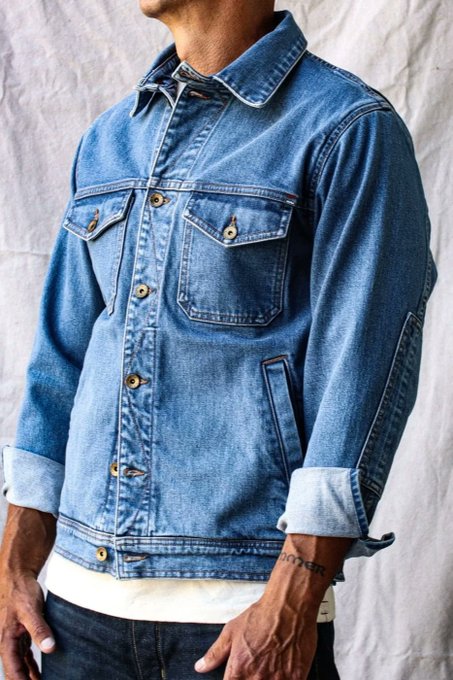 A man models a classic blue denim trucker jacket with two chest pockets and copper buttons, worn over a white t-shirt with sleeves rolled up.