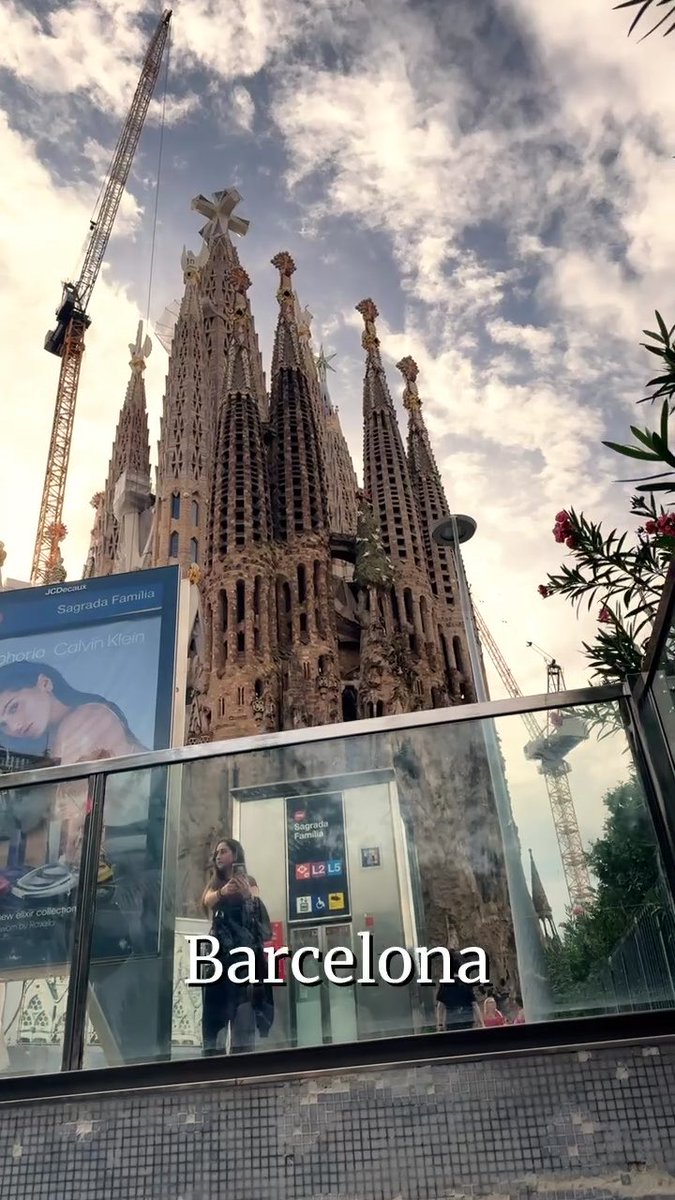 The Sagrada Família in Barcelona is shown towering over a glass-walled metro entrance, with a billboard for Calvin Klein to the left and the word 'Barcelona' overlaid on the image.