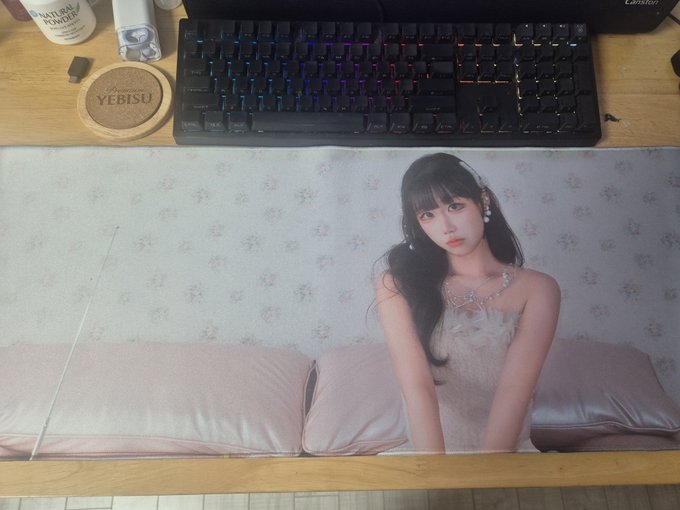 A large desk mat featuring a photo of a young woman in a white lace outfit sitting on a bed, placed in front of a backlit gaming keyboard.