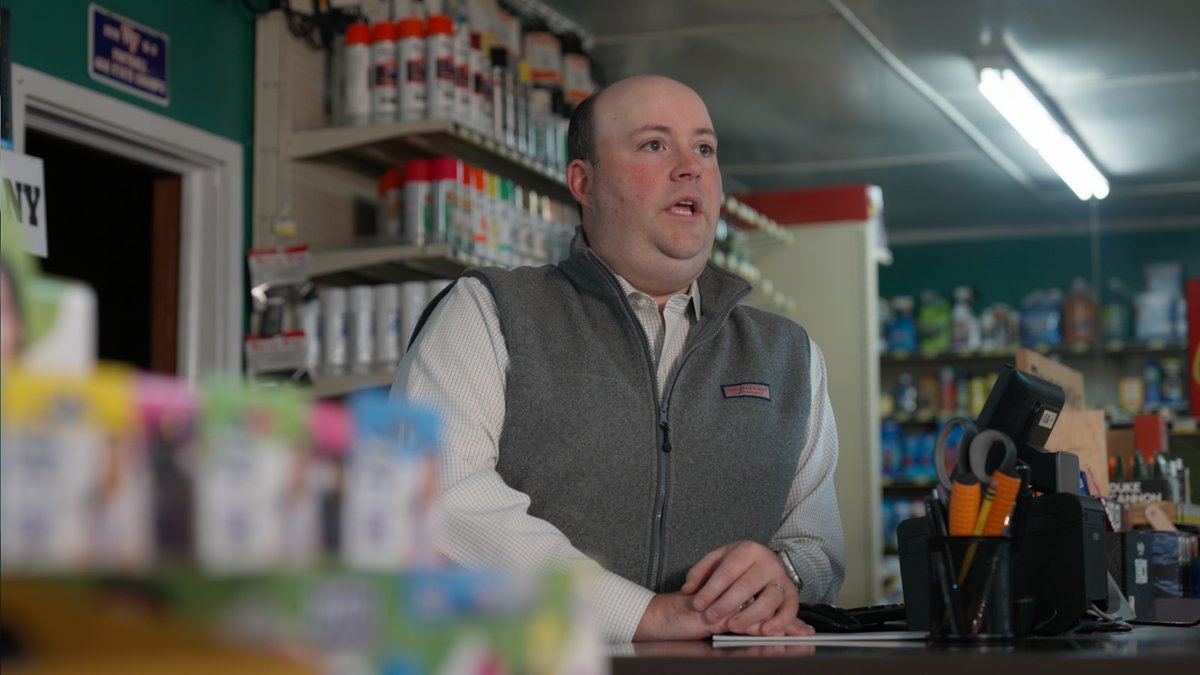 A photograph of a man wearing a grey fleece vest over a checkered shirt standing behind a counter in a store filled with various products on shelves.
