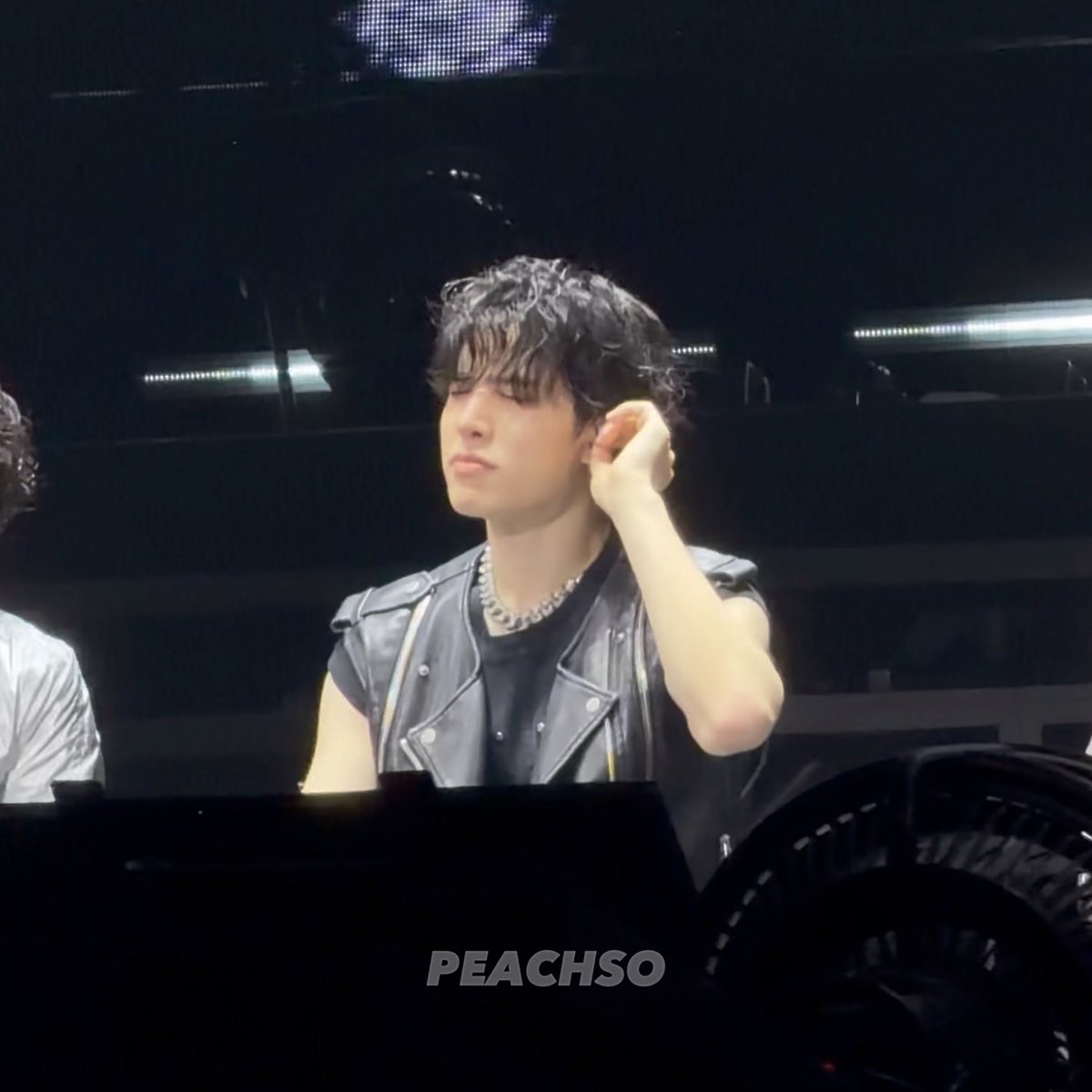 A young man with dark wavy hair wearing a black sleeveless leather jacket and silver chain necklace, appearing to be at a concert or event, with 'PEACHSO' watermark visible.