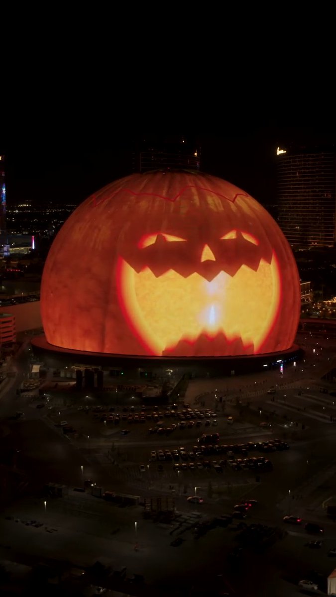 The Sphere in Las Vegas is illuminated as a giant glowing jack-o'-lantern at night, with a large grinning face projected across its massive dome as part of its Halloween-themed Exosphere show. Surrounding streets and parked cars are visible in the dim foreground, emphasizing the venue's enormous scale against the dark city skyline.