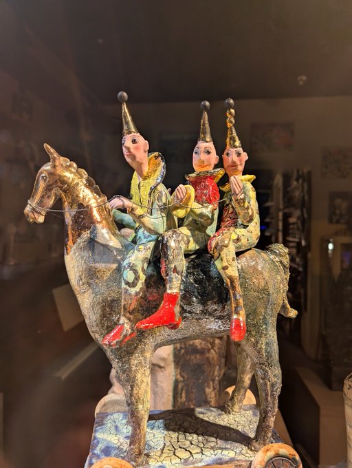 A colorful ceramic or paper-mâché folk art sculpture depicting three figures wearing pointed hats riding on the back of a horse, displayed in what appears to be a museum case.