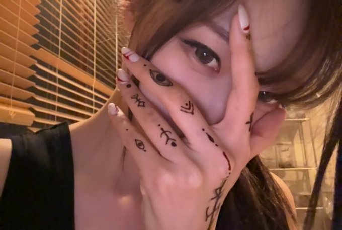 A close-up selfie of a young person with manicured nails covering part of their face, with several hand-drawn tattoos or markings including symbols and characters on their fingers.