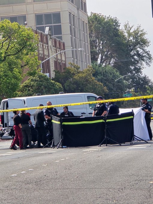 Police officers and emergency personnel respond to an incident on a city street, with yellow caution tape and a black privacy screen set up near white vans.