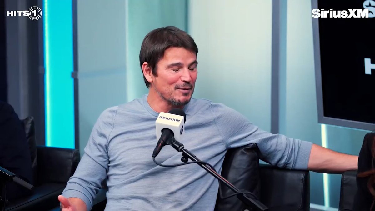 The image shows a man being interviewed on SiriusXM's HITS 1 radio show, speaking into a SiriusXM branded microphone. He appears to be mid-conversation, smiling slightly with his eyes closed. The SiriusXM and HITS 1 logos are visible in the upper corners of the studio setting, contextualizing the moment as the actor discusses his challenging experience on the set of "Verity."